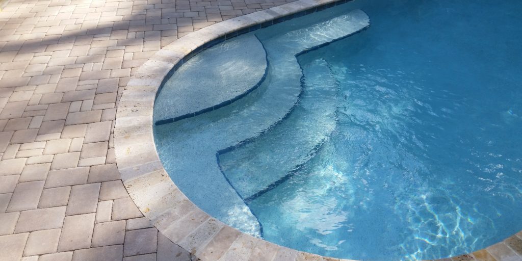 Free form swimming pool with travertine  coping stones and an interlocking concrete paver pool deck